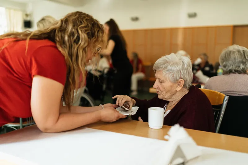 A carer offers afternoon tea to an older woman at a table