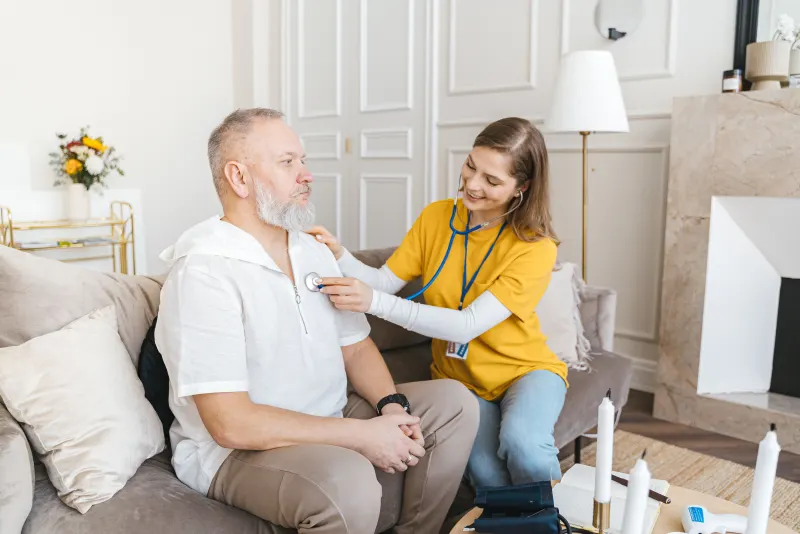 A nurse checks an older man's heart with a stethoscope in his living room